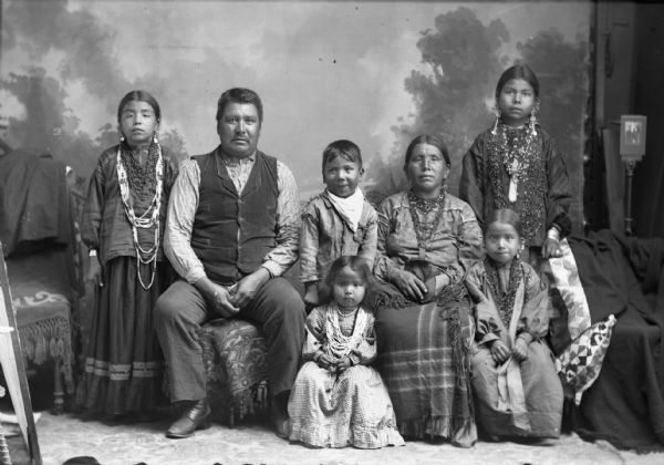 Studio portrait of a Ho-Chunk family, Jim White Bear, his wife Kate, and their children (four girls and a boy). Kate White Bear and the older girls are wearing degrees of regalia, and the remaining individuals are wearing contemporary dress.