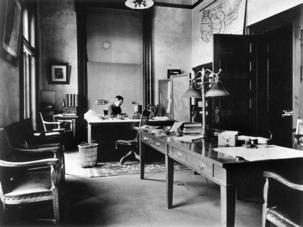 Interior view of the State Historical Society Library Superintendent's office with Reuben Gold Thwaites seated at his desk. There is a map of the United States on the wall next to the door.