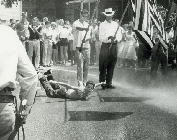 The police used fire hoses to control the angry crowds that gathered near Central High School during the integration crisis. There is a boy on the ground who has been drenched. Men stand next to him holding flags while people in the crowd are laughing.