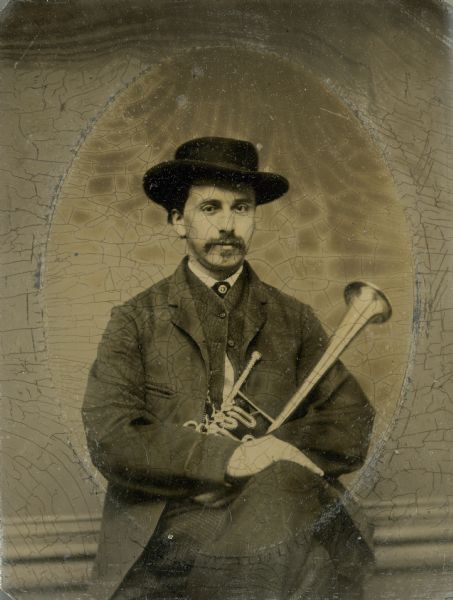 Studio portrait of E. (Edwin) O. Kimberley wearing a hat and holding a musical instrument in his lap, possibly a trumpet. Kimberley was leader of both the 3rd Wisconsin Infantry Regimental band and the 2nd Brigade, 4th Division, 15th AC.