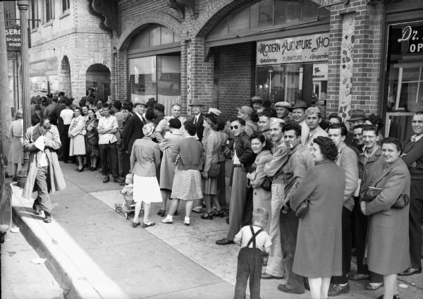 People lined up in front of Berg Sporting Goods Store, 2123 Atwood Avenue, to buy shotgun shells which were scarce due to World War II.  Also shows Modern Furniture Shop, 2119 Atwood Avenue, and Dr. R.F. Roman Optomitrist, 2117 Atwood Avenue.