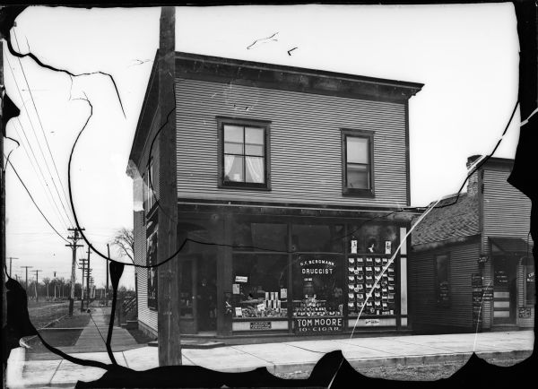 Exterior view of the shop of H.F. Bergmann, Druggist. There is a sign advertising Tom Moore Cigars in front of the building and a wooden sidewalk.