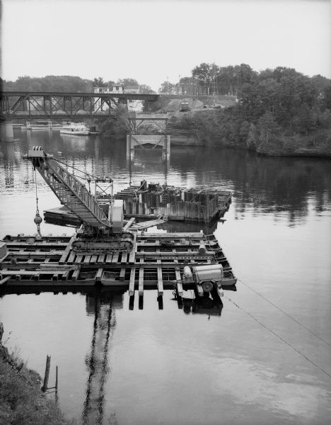 Construction equipment on a pontoon in the Wisconsin River during construction of the Wisconsin Dells bridge. Cars can be seen on the old railroad bridge in the background.