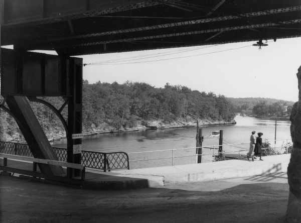Two women walking on the sidewalk along Broadway. Photograph taken from under the railroad bridge at Kilbourn (now Wisconsin Dells). The Wisconsin River is in the background.
