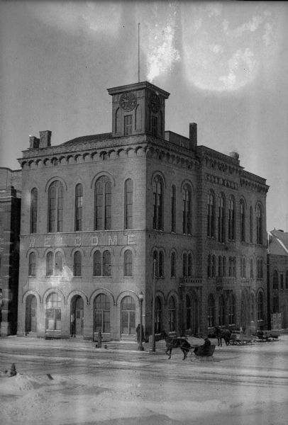 Exterior view of City Hall during winter with horse-drawn sleighs passing. A sign above the second story windows on the front of City Hall reads: "Welcome."