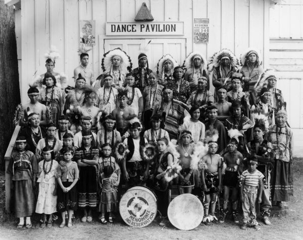 Peavey Falls group of dancers and musicians from the Menominee Indian Reservation in Wisconsin. The group is standing in front of a building with a sign that reads: "Dance Pavilion." A poster to the left of the door reads: "Menominee Indian Fair and Pageant, [Ke]shena, Wis. August 3-4-5 Pageant on August 4-5, 8 P.M."