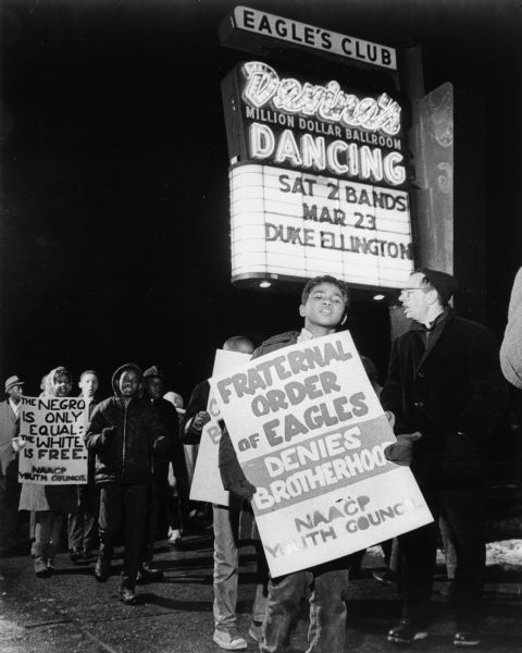 Father James Groppi and the NAACP Youth Council demonstrating outside the Eagles Club at night beneath a marquee advertising a Duke Ellington performance.