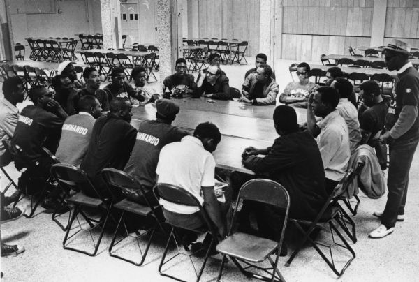 Father James Groppi meets around a large table with the NAACP Youth Council. Several of the men are wearing shirts with the word "Commando" on the back.