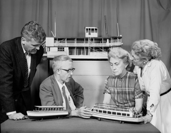 Contract Signing for Wisconsin Dells Minirama. The man on the left is a banker, the man with eyeglasses is Ted Saunders, and the women are Ruth Bennett Dyer and Miriam Bennett. There are model boats on the table in front of them, and another one is displayed behind the group.