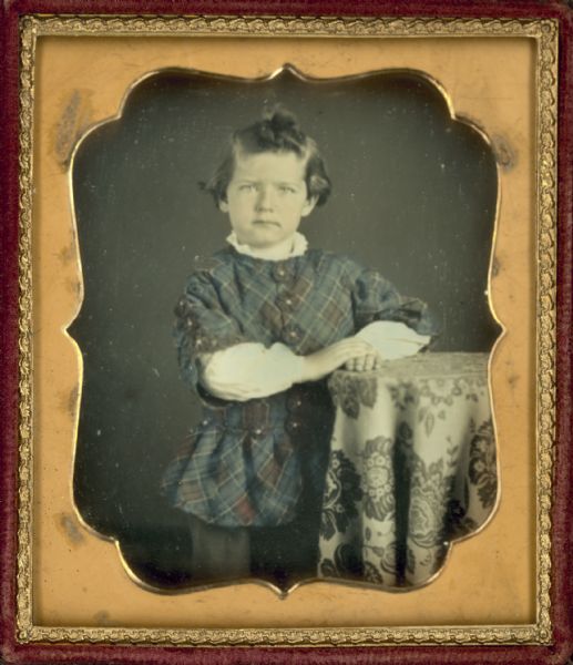 Sixth plate daguerreotype portrait of George Walter Oakley as a child standing next to a cloth-covered table with his both his hands resting on top of it, and wearing a white shirt under buttoned plaid tunic. Hand-coloring on cheeks and tunic.
