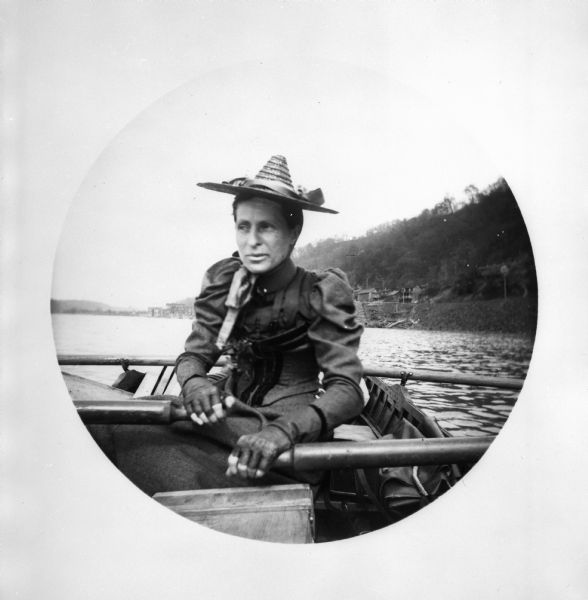 Jessie Thwaites wearing a fashionable dress and hat as she rows a boat on the Monongahela River.