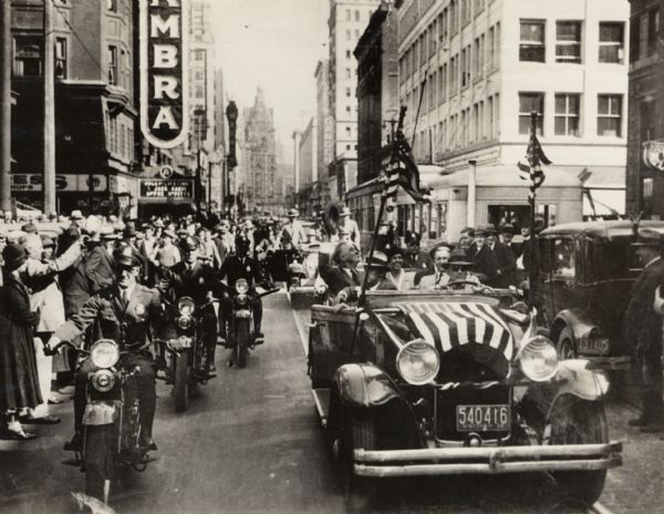 Franklin D. Roosevelt campaigning for the presidency in Milwaukee in 1932.  FDR is riding in a flag-draped open car and escorted by police on motorcycles.