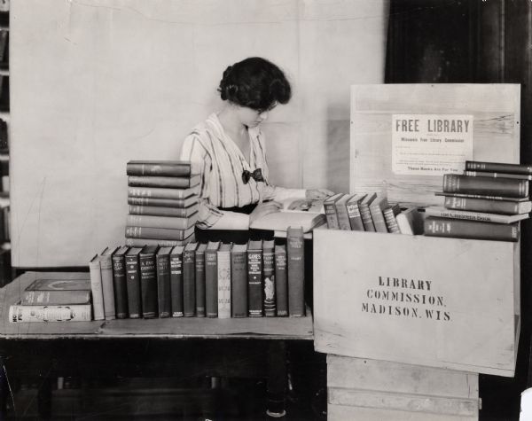 Woman, probably a librarian, paging through a book from a crate stamped "Wisconsin Free Library Commission, Madison, Wis." Several other books are stacked in and around the crate.