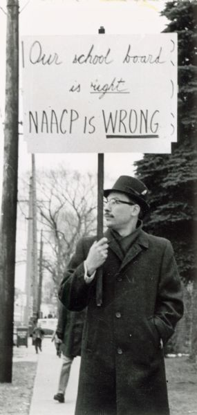 Man in a hat and long coat holding a sign opposing desegregation in schools. Possibly in Milwaukee.
