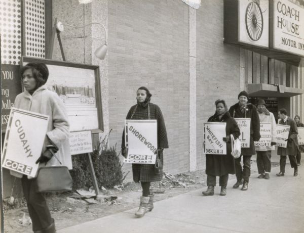 Marchers carrying signs bearing the names of Milwaukee suburbs during a CORE (Congress of Racial Equality) march for fair housing in Milwaukee. They are passing in front of the Coach House Motor Inn.