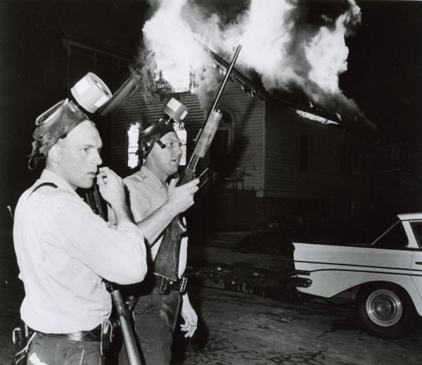 Freedom House in flames after Milwaukee Police fired tear gas into the building. Two armed officers stand in front of the burning building. Both officers have gas masks on their heads.