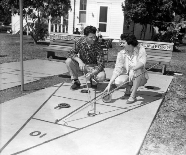 Andy Pafko, ballplayer for the Milwaukee Braves, and his wife playing shuffleboard. They are crouching in front of benches painted with advertising for Zoller Lumber Company.
