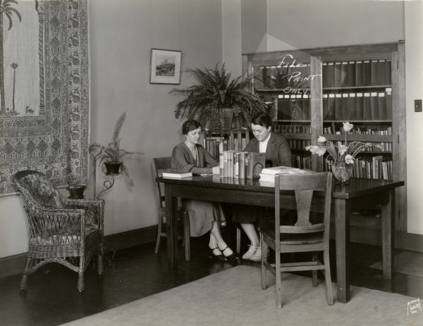 Two women studying at a table at Milwaukee-Downer College. There is a tapestry on one wall and a vase of flowers and several books on the table.