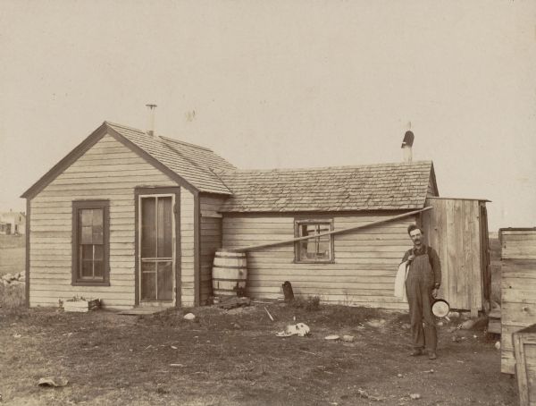 Man wearing coveralls standing in front of a small farm house. He has a towel over one arm and is holding a skillet in the other hand. There is a cat eating from a plate in front of the house as well.