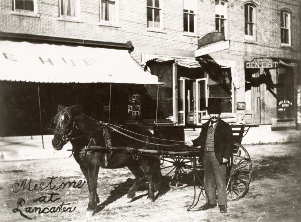Man in coat and top hat holding the harness of a horse attached to a buggy.