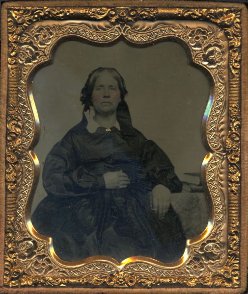 Quarter plate tintype/ferrotype of Lucetta Case (1808-1884). Three-quarter-length seated figure wearing dark dress with white collar, and ribbons in hair, with left elbow on table and right hand on torso. Hand coloring on cheeks, and gold details on pin at collar.