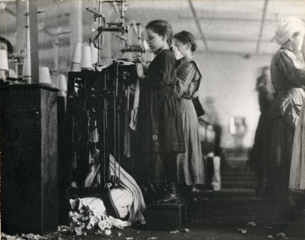 Young girls working with textile machinery. One girl is standing on a box to reach her work.