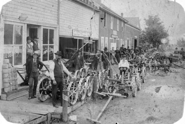 Large group of people posed in commercial area on a wooden sidewalk. Leigeois Bar, Implement Store, and Blacksmith Shop. Joseph Leigeois is shown in the forefront, John Leigeois has a moustache and is leaning on door entry. Rachel Leigeois is dressed in black. Children are sitting on agricultural machinery parked along the street.