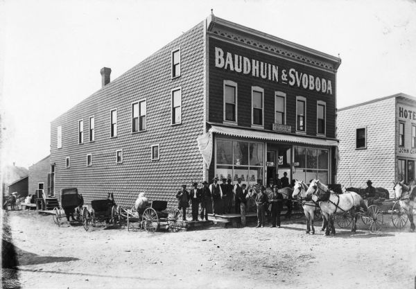 Men and boys posing with horses and horse-drawn vehicles in front of the Baudhuin and Svoboda store.  There is a Post Office sign on the building.