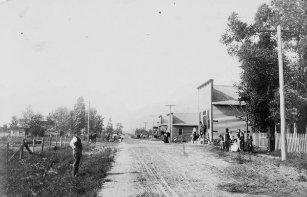 People are standing and sitting along the dirt road. There are a few framed buildings, a woman holding a horse, and a group of people in front of a fenced-in yard.