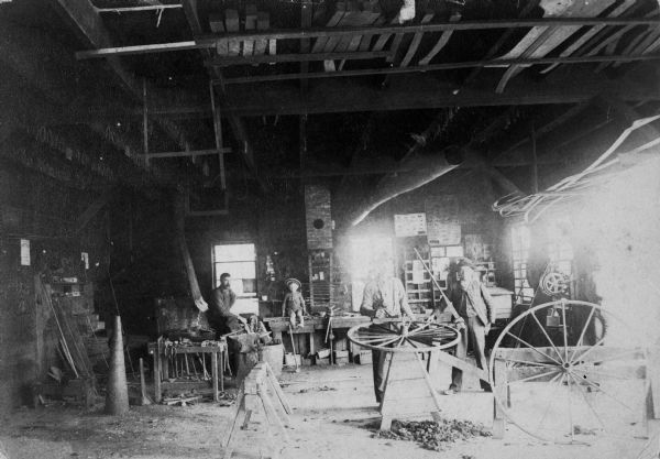 Interior of shop. John Liegeois is sitting on bench, Louis is at wheel. A young child with a hat sits on a bench, and another man is also standing near the wheel.
