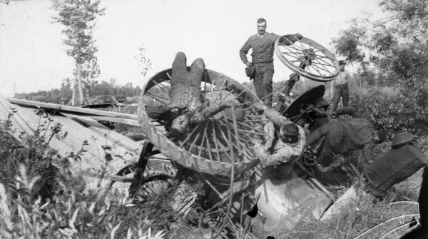 Group of men staging an accident. Two men stand nearby watching as three other men pose on the wheels of an overturned vehicle.