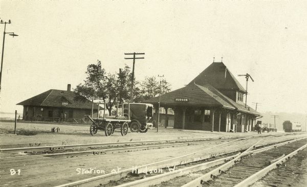 View across railroad tracks towards the railroad station in Hudson. There is a truck and a wagon parked alongside the tracks. A railroad car is parked just beyond the station on the right.
