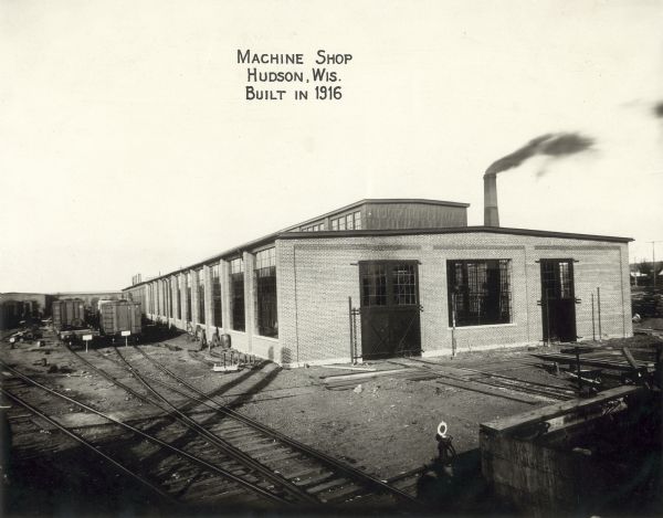 Slightly elevated view of a machine shop in Hudson, which was built in 1916. There are railroad tracks and railroad cars along the left side of the building. A smokestack is behind the building on the right.