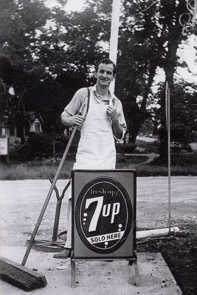 Future District Court Judge William Sachtjen, wearing an apron and holding a broom. He is standing behind a 7-Up sign. Sachtjen worked at Droster's Grocery Store while he was in school.
