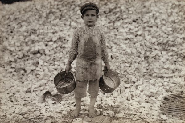 Child laborer standing barefoot near a pile of shells and holding a strainer in each hand.