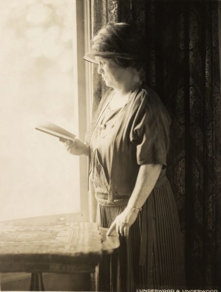 Belle Case La Follette standing at a window and reading.