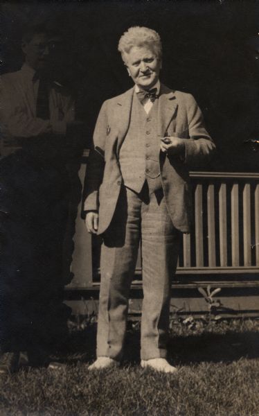 Robert M. La Follette standing and holding a pipe. Philip La Follette stands in the background in the shadows.