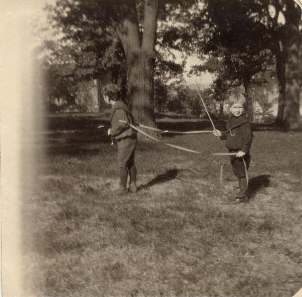 Young Robert La Follette, Jr. and Philip La Follette playing in the yard. Robert wears a harness and Phil holds a crop.