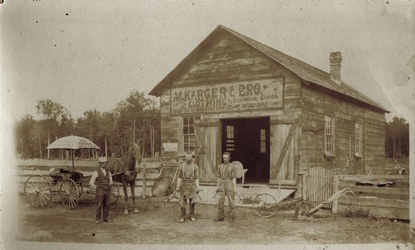 Three men and a horse standing in front of a blacksmith shop which has a sign above the doorway advertising M. Karger and Brothers Clothing and Furnishing Goods store. Standing on the right is Rob Schwager, and in the center is Fred Paul (1855-1931), a descendant of the Reinhold Paul family from Edgar, Wisconsin. There is another man on the left wearing a hat.