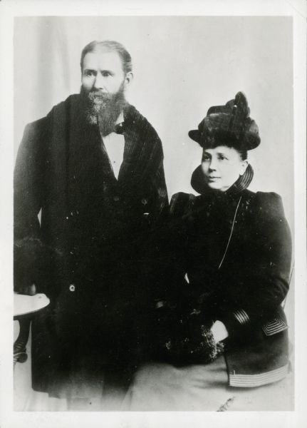 Studio portrait of Jeremiah Curtin and Alma Cardell Curtin. Both are wearing coats and she is wearing a hat.