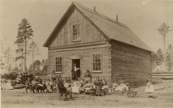 Native American children, presumably students, and adults pose outside an Indian school. The location is not verified, but may be in the Hayward vicinity.