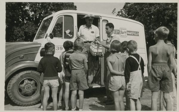 A group of children gather around a milk truck as the milkman emerges with bottles of milk.