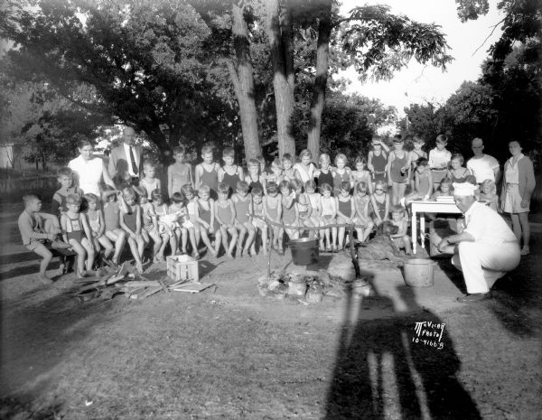 Kiddie Camp children around campfire, watching frankfurters roast in a pot over the fire, tended by Oswald B. Neesvig, president of Madison Packing Co. Most of the children are wearing bathing suits.