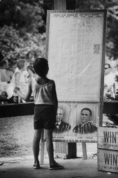 Young girl standing in front of a large document of family history displayed on a board. There is a large group of people in the background.