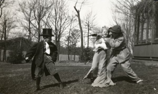 Philp La Follette, Mary La Follette, and Robert La Follette, Jr. playing in costume in the front yard of their home. Phil wears a top hat and long coat. Robert appears to be playing the villain holding Mary hostage with guns.
