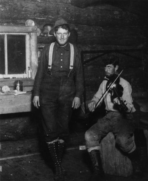 Two men in a lumber camp bunkhouse. The man on the right is sitting on a stump playing a fiddle, while the man on the left is dancing.