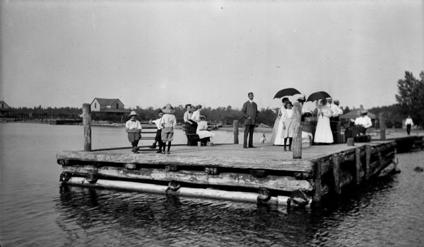 John Pitt Dean standing at the center of Mission Dock surrounded by people, some of them holding umbrellas. There is pile of luggage on the dock.