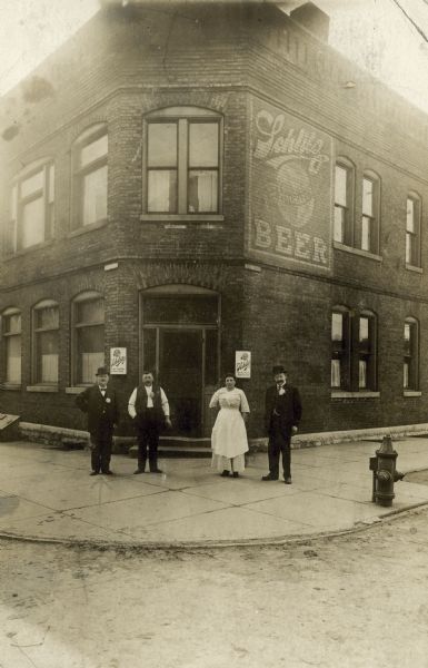 Exterior view of the bar owned by Rudolph Steinbacher at the corner of 16th Street and St. Paul Avenue.  A large sign for Schlitz beer is painted on the wall and four people, perhaps the owner and employees, are standing in front.