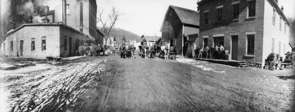 Panoramic view showing, on the left, the bottling plant and the stables, and on the right, the Potosi Brewery itself. Groups of men are standing on the sidewalks, and three men are driving horse-drawn vehicles down the street.