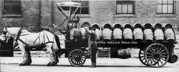 Man standing in front of horse-drawn wagon loaded with barrels of beer from the Miller Brewery. The sign near the wagon seat reads: "No.2" and the sign painted on the wagon reads: "Drink Miller High Life."